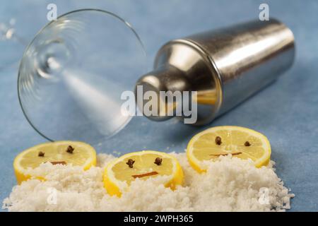 tranches de citron avec clous de girofle et cannelle formant un visage souriant sur un tas de sel à côté d'un shaker à cocktail et un verre de cristal avec fond bleu Banque D'Images