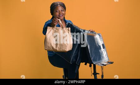 Jeune femme noire en bonne santé donne les pouces tout en livrant des aliments à emporter assurant la satisfaction du client. Livreur afro-américain fournissant une alternative écologique à dîner dans le quartier. Banque D'Images