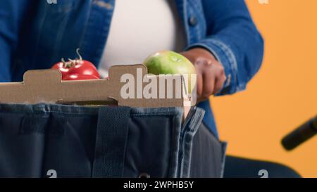 Courrier féminin avec origine ethnique afro-américaine déplaçant délicatement le panier de produits frais du sac de livraison de nourriture. Femme noire enthousiaste sur la bicyclette tenant la boîte de légumes. Gros plan portable. Banque D'Images