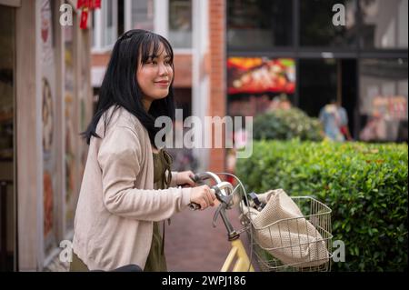 Une belle femme asiatique positive dans une robe mignonne marche et pousse son vélo dans la ville, explorant la ville par une journée ensoleillée. Banque D'Images