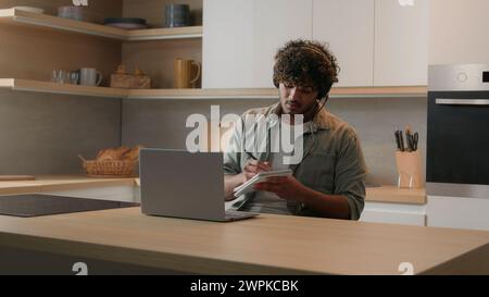 Homme indien dans des écouteurs assis à la table dans la cuisine domestique regardant la vidéo éducative appel leçon de classe sur ordinateur portable écrivant des notes arabes Banque D'Images