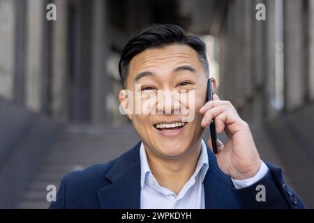 Joyeux homme d'affaires asiatique vêtu de façon décontractée d'un costume faisant un appel téléphonique heureux tout en se tenant debout à l'extérieur. Banque D'Images