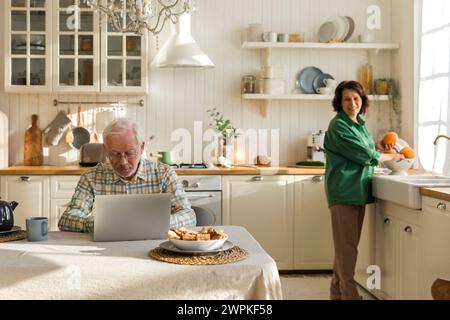 l'homme âgé dans des lunettes utilise un ordinateur portable dans la cuisine Banque D'Images