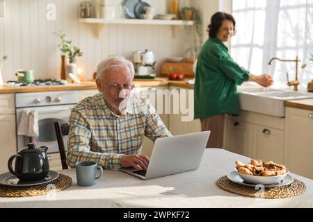 homme âgé aux cheveux gris dans les lunettes utilise l'écran d'ordinateur portable dans la cuisine Banque D'Images