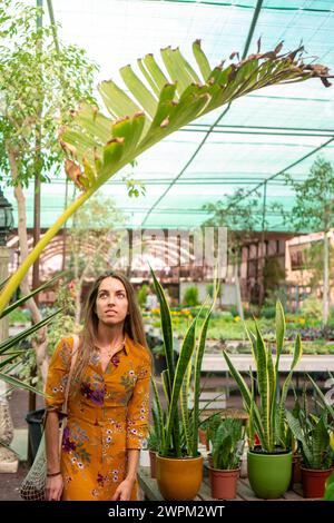 Une femme portant une robe jaune se tient gracieusement dans une serre luxuriante, entourée de diverses plantes et fleurs vibrantes. Banque D'Images