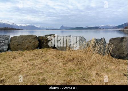 Un bord de mer tranquille avec de grands rochers bordant le rivage, des montagnes lointaines visibles au-delà de l'eau placide, créant une atmosphère sereine. Banque D'Images