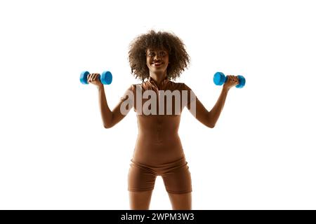 Femme afro-américaine souriante soulevant des haltères bleus, vêtue d'un costume d'entraînement marron sur fond blanc de studio. Banque D'Images