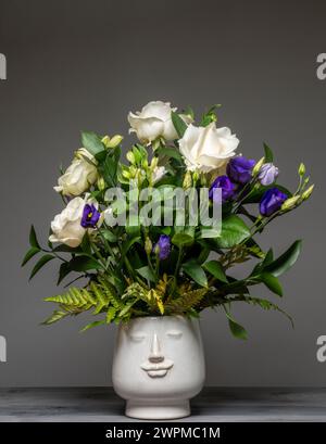 Arrangement floral de roses blanches, lisianthus pourpre et feuillage disposés dans un vase de visage en céramique blanche. Banque D'Images