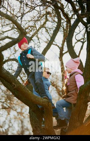Heureux, joyeux enfants, garçons et filles, en tenue d'hiver grimpant à l'arbre, profitant de l'aventure ensoleillée en plein air. Banque D'Images