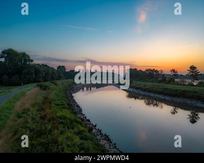 Cette image tranquille capture la première lumière de l'aube se reflétant sur les eaux calmes d'un canal, avec les teintes pastel du ciel du matin reflétées sur sa surface. Un sentier longe le canal, invitant à une promenade ou à un vélo tôt le matin. Le canal est flanqué de remblais verts et d'arbres clairsemés, dont les silhouettes deviennent visibles dans la douce lumière du matin. Cette scène transmet un sentiment de solitude paisible et la beauté des matins tôt dans un cadre pastoral. Le calme de l'eau et le calme de la pause de la journée en font une représentation idyllique de l'élégance simple foun Banque D'Images