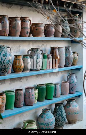 Des pièces de poterie astucieusement conçues ornent les étagères bleues, présentant une riche variété de glaçures et de motifs peints à la main. Chaque pièce fait écho à l'art traditionnel de la céramique. Banque D'Images