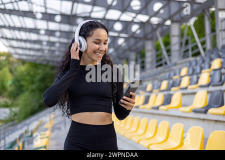 Une femme souriante écoute de la musique à travers des écouteurs blancs, en utilisant son smartphone dans un coin salon vide du stade. Banque D'Images