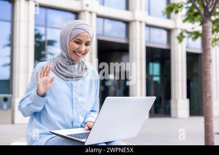 Joyeuse dame musulmane portant un hijab s'engageant dans un chat vidéo, agitant à la webcam avec un sourire joyeux à l'extérieur. Banque D'Images