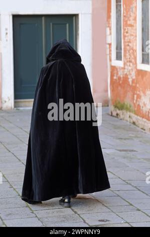 figure dans une capuche marche à travers une allée de la ville portant un tabard noir usé comme un manteau Banque D'Images