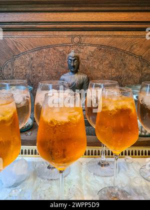 Plusieurs verres de boisson gazeuse glacée et des glaçons sont flous au premier plan devant un mur en bois avec une figure en bois de Bouddha Banque D'Images