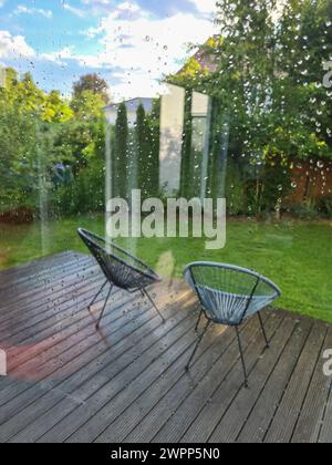 Vue à travers la vitre avec des gouttes de pluie sur la terrasse humide avec deux chaises de jardin modernes Banque D'Images