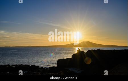 Image de silhouette d'un homme pêchant sur les rochers. Lever du soleil sur l'île de Rangitoto. Takapuna Beach. Auckland. Banque D'Images