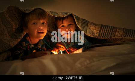 Couverture, lampe de poche et les enfants la nuit avec bonheur en portrait avec dessin dans un livre. Amis, détendez-vous et dessinez sur ordinateur portable dans l'obscurité avec la lumière Banque D'Images