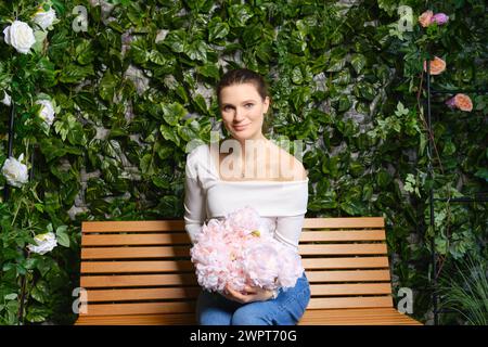 Femme mignonne assise sur un banc en bois tenant un bouquet de délicates fleurs roses sur ses genoux Banque D'Images