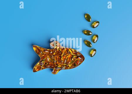 Boîte en forme de poisson remplie de pilules elliptiques orange d'huile de poisson et de quelques capsules formant des bulles sur fond bleu. Suppléments d'acides gras oméga-3 Banque D'Images