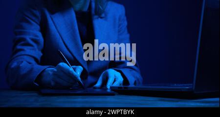 Femme d'affaires travaillant sur une tablette graphique et un ordinateur portable dans l'obscurité et éclairé par l'écran. Pas de face. Concentrez-vous sur les mains. Correction des couleurs de gris. Banque D'Images