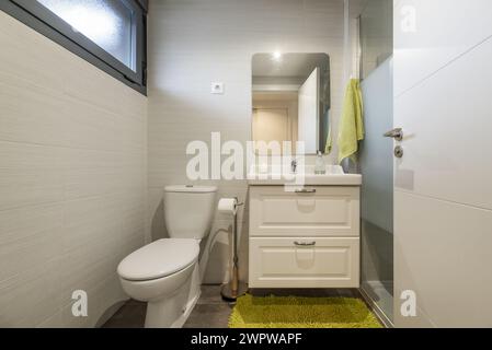 Une salle de bain conventionnelle avec armoire en bois blanc, évier en porcelaine d'une seule pièce au-dessus de la commode, miroir sans cadre, tapis vert et verre trempé Banque D'Images