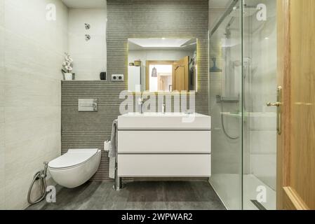 salle de bains avec toilettes en porcelaine blanche avec tiroirs et cabine de douche avec écrans en verre trempé, armoire à tiroirs blanche sous le miroir et bois de chêne do Banque D'Images