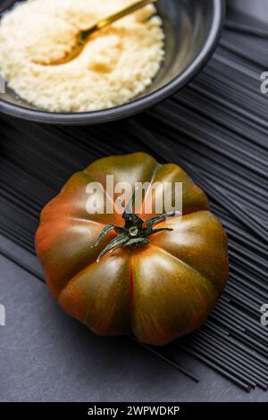 Une succulente tomate raf mûre sur un tas de pâtes noires et un bol de fromage râpé Banque D'Images