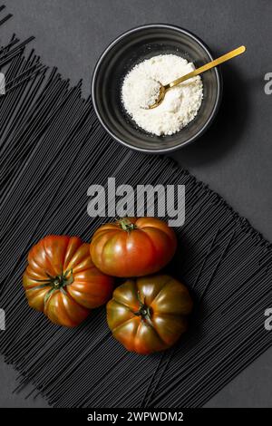 Un trio de succulentes tomates Raf mûres sur un monticule de pâtes noires à côté d'un bol plein de fromage râpé sur une surface lisse et noire Banque D'Images