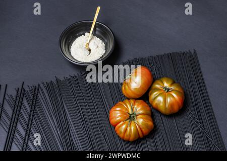 Un trio de succulentes tomates Raf mûres sur des spaghettis teints en noir, le tout sur une surface noire lisse à côté d'un bol noir rempli de fromage râpé Banque D'Images