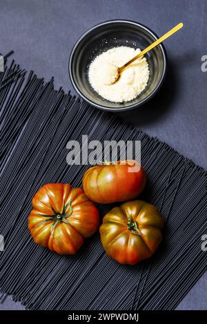 Un trio de succulentes tomates Raf mûres sur des spaghettis teints en noir, à côté d'un bol noir rempli de fromage râpé avec une cuillère dorée Banque D'Images