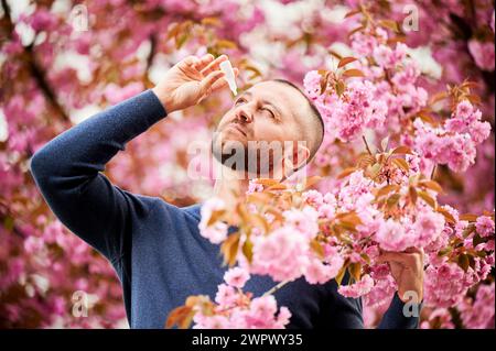 Homme allergique appliquant des gouttes pour les yeux médicaux, souffrant d'allergie saisonnière au printemps dans le jardin fleuri. Bel homme traitant les yeux devant un arbre en fleurs à l'extérieur. Concept d'allergie printanière. Banque D'Images