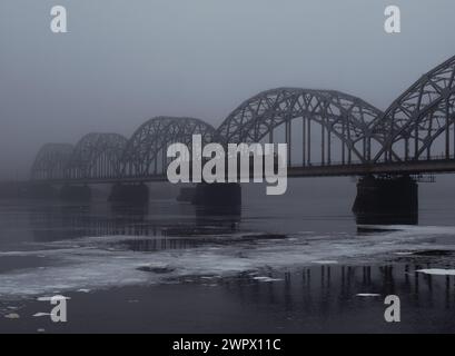 Pont ferroviaire de Riga sur la rivière Daugava brumeuse avec train traversant le pont 28.02.2024 Banque D'Images