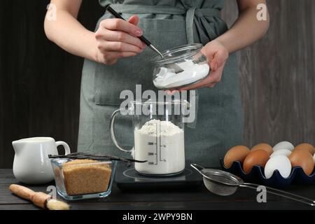 Femme ajoutant de la levure chimique dans la tasse à mesurer à la table en bois noir, gros plan Banque D'Images