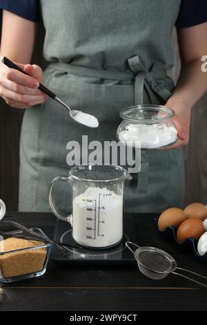 Femme ajoutant de la levure chimique dans la tasse à mesurer à la table en bois noir, gros plan Banque D'Images