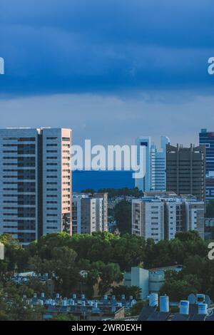 Paysage urbain côtier : tours modernes, verts verdoyants et mer lointaine sous ciel bleu Banque D'Images