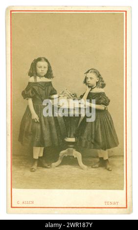 Carte de visite victorienne originale (carte de visite ou CDV) de deux jeunes sœurs galloises sérieuses, filles victoriennes, enfants victoriens, portant des robes de style crinoline à la mode, des manches bouffantes et des bottes, avec de longs cheveux sertis tenus avec un arc, des pendentifs et des jouets de maintien éventuellement. Du studio photographique de C. Allen, Tenby au pays de Galles. Circa 1864 de manches bouffantes, coiffure, robe. Banque D'Images