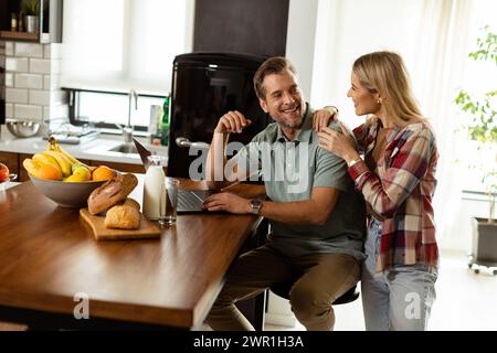 Un couple joyeux profite d'un moment léger dans leur cuisine ensoleillée, travaillant sur ordinateur portable entouré d'un petit déjeuner sain Banque D'Images