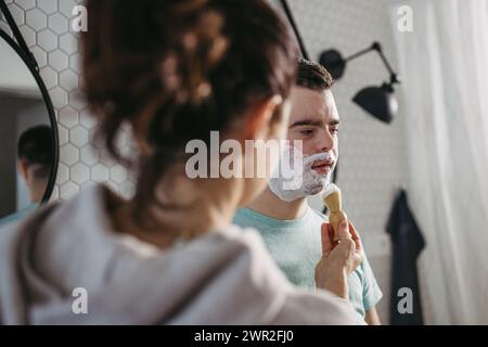 Mère appliquant de la mousse à raser sur le visage de son fils. Jeune homme trisomique apprenant à se raser. Banque D'Images