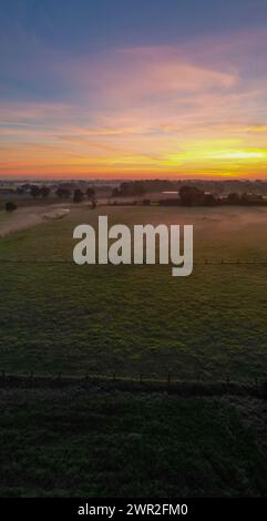 Capturée à l'aube, cette image incarne la beauté sereine d'une scène de campagne baignée par la douce et chaude lueur du lever du soleil. L'horizon est peint avec une palette riche d'oranges, de roses et de violets au fur et à mesure que la journée commence à se dérouler. Ci-dessous, une vaste étendue de prairies ouvertes est légèrement embrassée par la lumière du matin, soulignant le paysage texturé et la silhouette nette d'une clôture qui délimite le champ. La tranquillité du cadre est palpable, offrant un moment de calme et de connexion à la vie pastorale. Aube pastorale : lever de soleil serein sur une campagne tranquille. Q élevé Banque D'Images