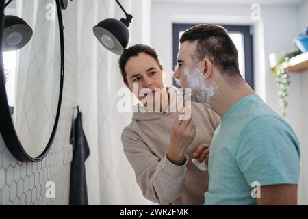 Mère appliquant de la mousse à raser sur le visage de son fils. Jeune homme trisomique apprenant à se raser. Banque D'Images