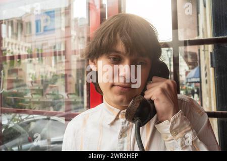 Jeune homme réfléchi dans la chemise rayée claire fait appel de la vieille cabine téléphonique publique, reflets de la ville en arrière-plan. Banque D'Images