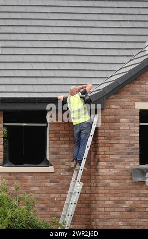 Un ouvrier de la construction ou un constructeur sur une échelle adaptant des gouttières au toit d'une maison neuve en construction Banque D'Images