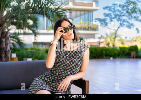 Une belle femme est assise sur un canapé dehors tout en enlevant ses lunettes de soleil Banque D'Images
