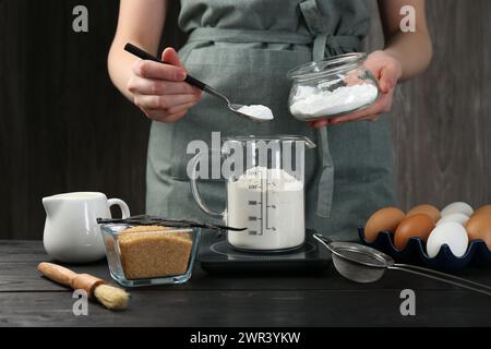 Femme ajoutant de la levure chimique dans la tasse à mesurer à la table en bois noir, gros plan Banque D'Images