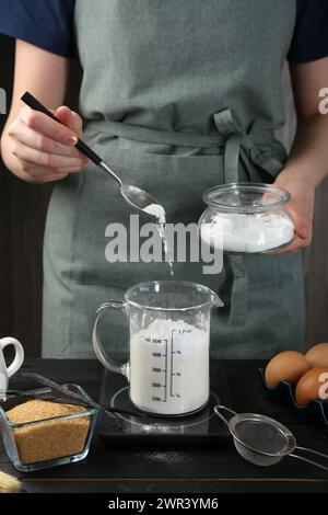 Femme ajoutant de la levure chimique dans la tasse à mesurer à la table en bois noir, gros plan Banque D'Images