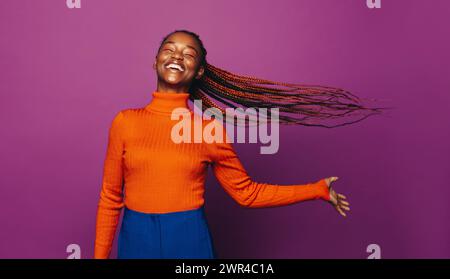 Femme africaine dans la vingtaine avec des tresses bicolores élégantes se tient dans un studio, souriante. Elle porte des vêtements décontractés, un Jean et un col polo, célébrant la vie wi Banque D'Images