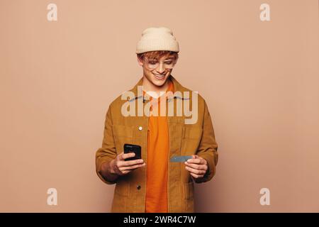 Jeune homme joyeux dans des vêtements décontractés se tient dans un studio avec une toile de fond pêche. Il détient un smartphone et une carte de crédit, en utilisant une application bancaire mobile pour e. Banque D'Images
