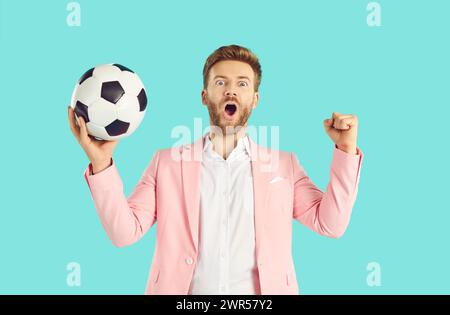 Jeune homme excité en costume rose tenant le football avec un visage choqué surpris sur fond bleu. Banque D'Images