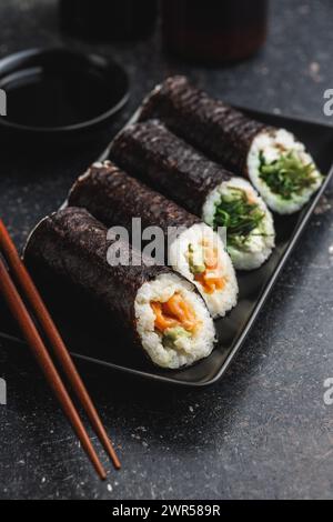 Une assiette noire avec des morceaux de sushi avec des baguettes et un bol de sauce soja. Le sushi montre une variété de couleurs, de textures et de garnitures, showc Banque D'Images